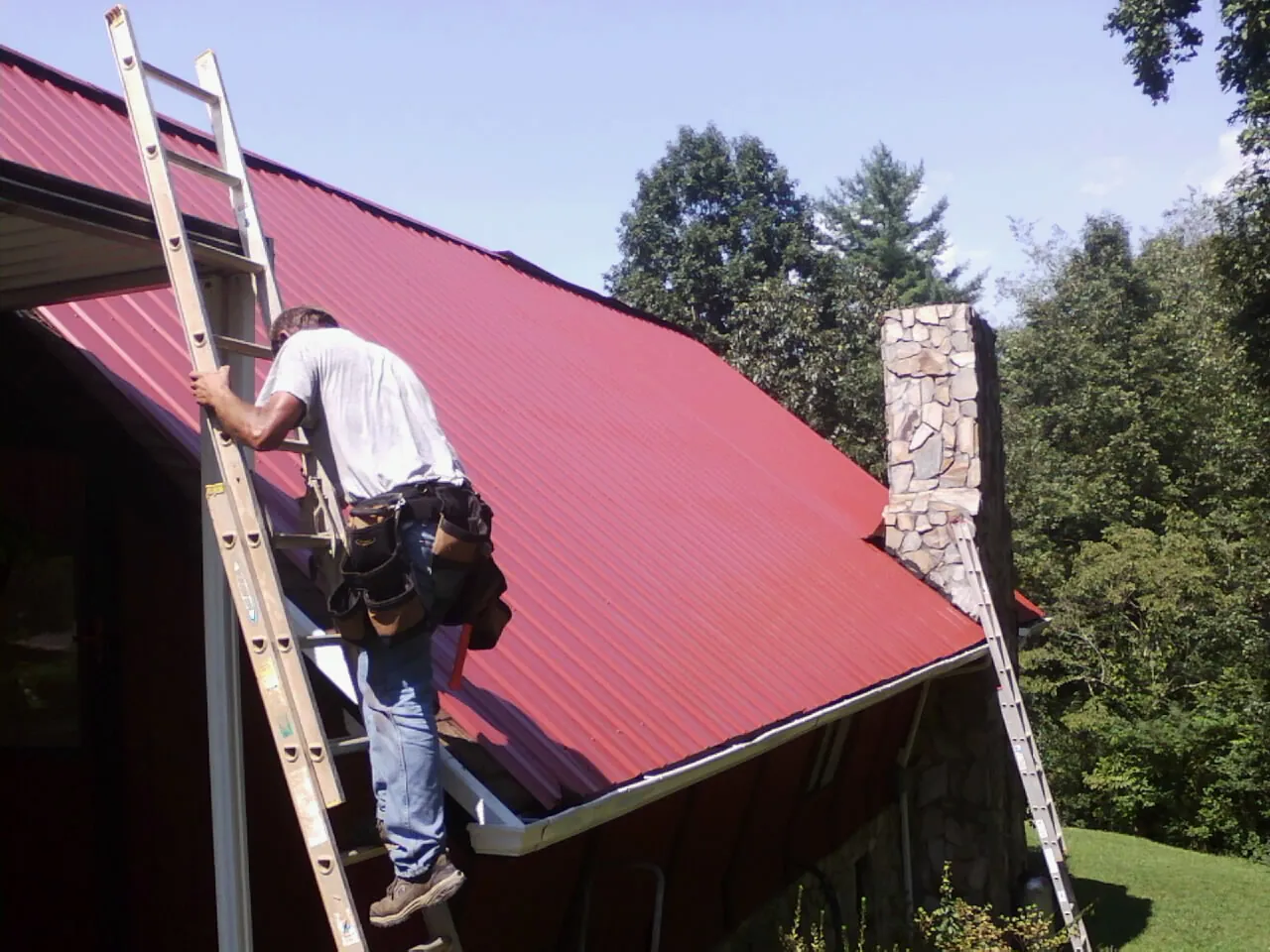 Roofer on a ladder beside a red metal roof and stone chimney — Soffit & Fascia Repair by Roofing Brothers in Oregon City